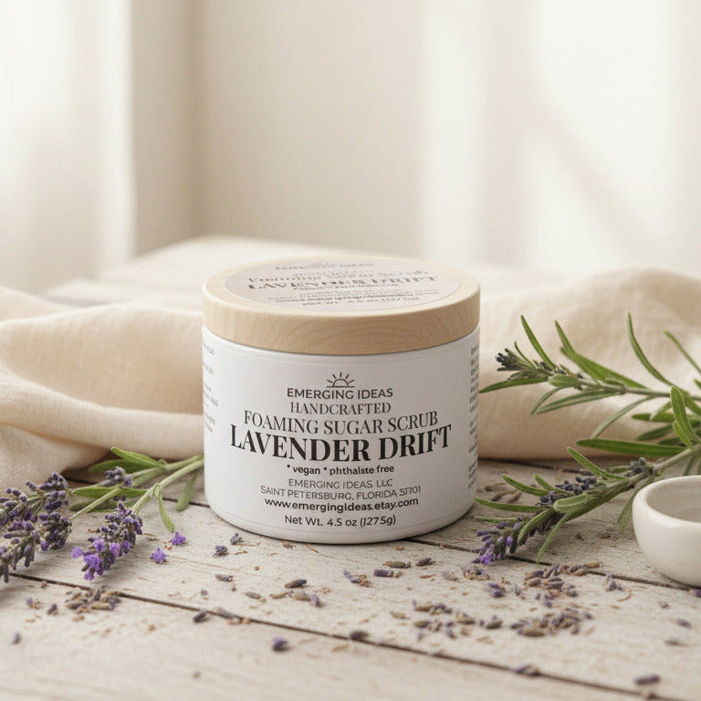 A jar of Emerging Ideas Handcrafted Lavender Drift Foaming Sugar Scrub sits on a rustic wooden tray. The product has a lavender label and a light wood lid, with the brand name 'Emerging Ideas' clearly visible.