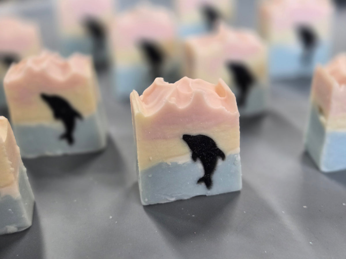 Layered handmade soap bars with pastel pink, yellow, and  blue sunset design, each featuring a black silhouette of a jumping dolphin.